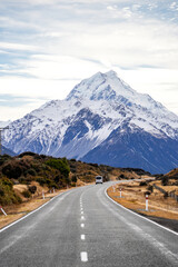 A stunning view of a snow-covered mountain towering above the landscape. A long road stretches into the distance, lined with dry vegetation. The image captures the vastness and natural beauty of this