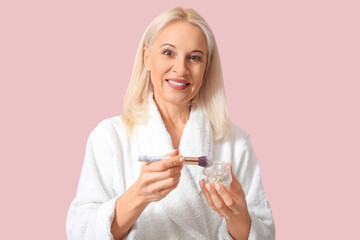 Mature woman in bathrobe with makeup powder and brush on color background