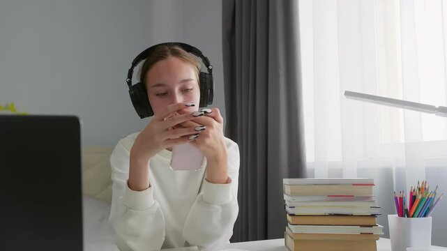 Teenage student wearing headphones is using a smartphone while seated at a desk with books, a laptop, and school supplies, suggesting a struggle with distractions and procrastination