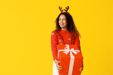 Happy young African-American woman in Christmas sweater and deer horns with gift box on yellow background