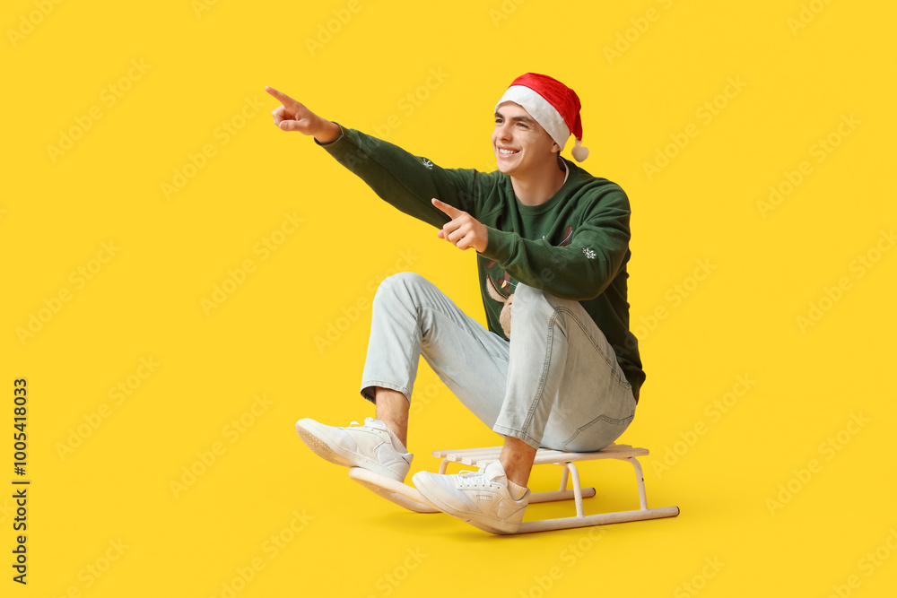 Poster Happy young man in Christmas sweater and Santa hat on sledge against yellow background