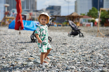 A toddler in a cute outfit and a sunhat walks along a rocky beach near the ocean as waves crash...