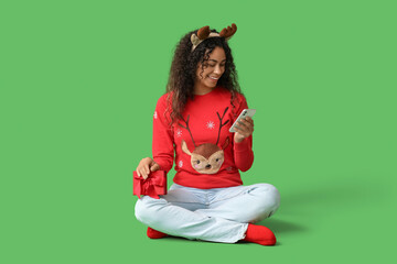 Beautiful young African-American woman in reindeer horns with mobile phone and Christmas gift box sitting on green background