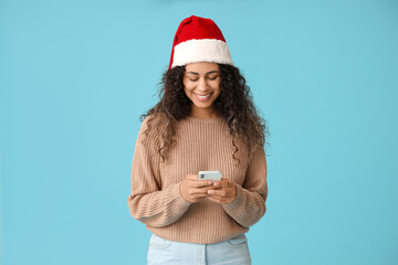 Beautiful young African-American woman in Santa hat with mobile phone on blue background. Christmas celebration