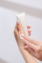 Woman's hand applying hand cream out of a mockup of white tube of cream. Unlabeled packaging for cosmetics in sunlight on white background. Concept of skincare. Harsh shadows