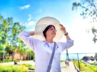 A woman is playfully adjusting her large sun hat while standing in a vibrant park filled with trees under a clear blue sky