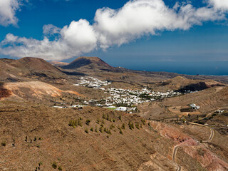 The volcanic landscape of Lanzarote presents a rugged terrain with reddish-brown hills, sparse vegetation, and winding roads. White houses contrast against the barren earth, with the ocean visible in 
