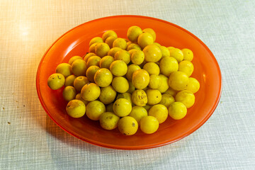 Heap of yellow tkemali fruits on a red plastic plate on light table. Prunus cerasifera, cherry plum, myrobalan plum