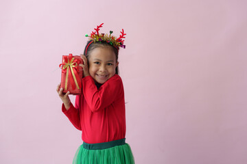 Portrait on a pink background of a cute little girl in a headband holding a happy red gift box with a beautiful green bow in various verses. Christmas Eve New Year and Winter holidays.