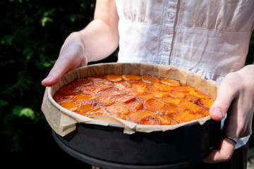 Close-up of a faceless girl holding a freshly baked apricot tart in a round pan, set against a natural, outdoor background. tart has a golden, glazed finish. Blur and selective focus