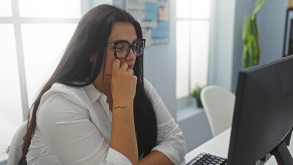 Young, hispanic, plus-size woman working at desktop computer in a modern office setting with white furniture and natural light from large windows.