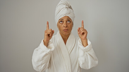 Middle-aged hispanic woman in a bathrobe with a towel on her head, pointing upwards against an isolated white background