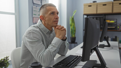 Man working alone in office, thinking, focused on computer, under natural light, indoor setting, workplace, office environment with plant and storage boxes in background using desktop