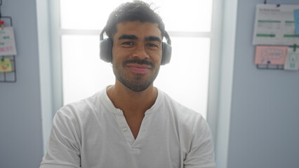 Handsome hispanic man wearing headphones in a bright office space with motivational notes on the wall