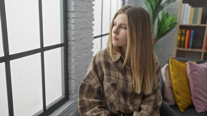 A thoughtful young blonde woman in a plaid shirt sits indoors near a window, looking away in a modern living room.