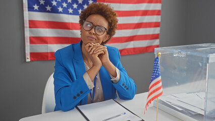 Thoughtful african american woman in blue sits by a ballot box, a us flag in the background,...
