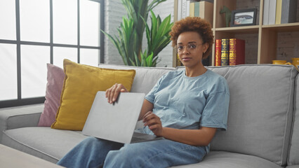 African american woman holding laptop, sitting on couch in a modern house interior, looking relaxed and confident.