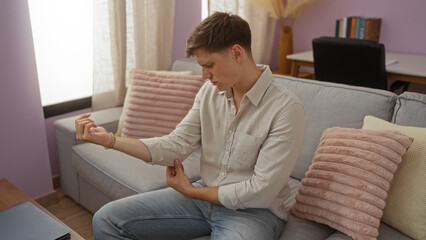 Fototapeta premium Young man in a living room holding his elbow and seeming to be in discomfort, surrounded by cozy interior elements like a couch and cushions