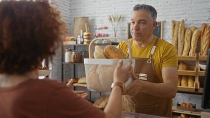 Man in bakery serving bread to woman customer indoors with shelves of various baked goods in the background