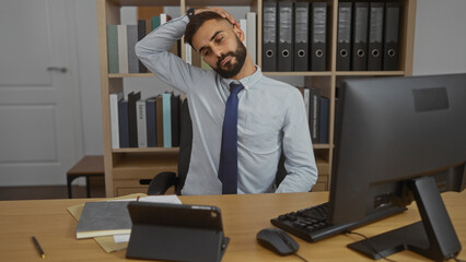 Young hispanic man with a beard working in an office wearing a tie, stretching his neck while sitting at a desk with a computer and bookshelves in the background.