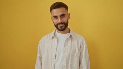 Handsome hispanic man with a beard standing over an isolated yellow background in a portrait style photo