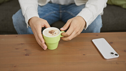 Man in casual wear enjoying coffee indoors, with smartphone on wooden surface nearby, depicting relaxation and technology.