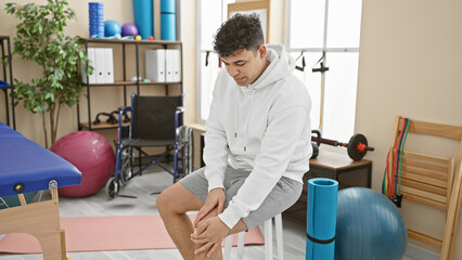 A young man in a white hoodie sits feeling knee pain in a modern physiotherapy clinic.