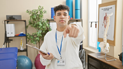 A young bearded man in a white shirt pointing at camera in a physiotherapy clinic with medical equipment.