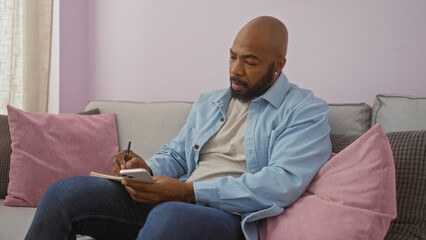 Young man sitting in a comfortable living room, using a smartphone and writing in a notebook