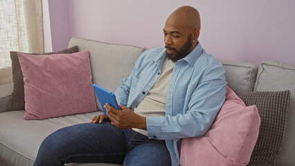 Young african american man with a beard sitting in a cozy living room on a couch using a tablet indoors at home