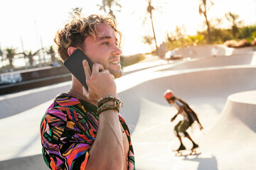 side view of positive millennial curly haired guy in stylish outfit talking on mobile phone while standing near concrete ramp in outdoor skatepark in summer