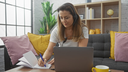 A focused hispanic woman wearing headphones takes notes at home, indicating remote work or study.