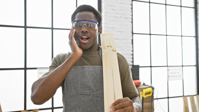 Surprised african man with safety goggles holding lumber in a bright woodworking workshop.