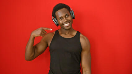 A smiling young african american man points to himself while wearing headphones against a red background.