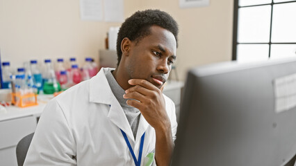 Fototapeta premium A contemplative young african man wearing a lab coat in a laboratory looking at a computer screen, representing healthcare professionals.