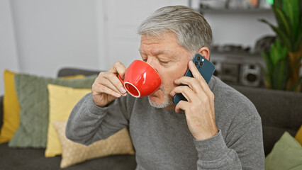 A middle-aged man multitasks, sipping from a red mug while talking on a smartphone in a cozy living...
