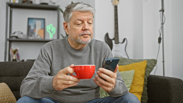 Mature man using smartphone and holding coffee mug in a modern living room.
