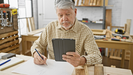 A mature man uses a tablet while drafting plans in a well-equipped carpentry workshop.