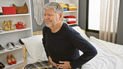 Mature grey-haired man with pain sitting on a bed inside a bedroom looking uncomfortable