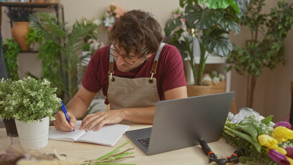 A focused hispanic man with a beard writes in a notebook at a flower shop, surrounded by plants and a laptop.