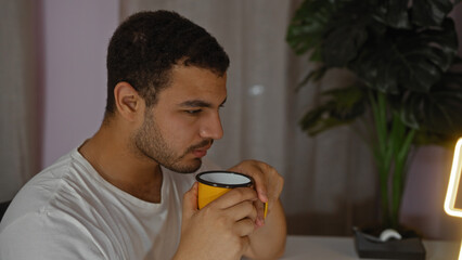 Young hispanic man enjoying a hot drink in a cozy living room, illuminated by warm lighting