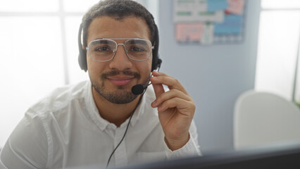 Young hispanic man wearing glasses and a headset smiling in an office environment indoors implying...