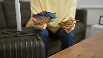 Hispanic woman counts swiss francs before travel in her living room, next to a luggage.