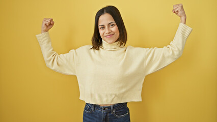 Young hispanic woman flexing arms against isolated yellow background, showing strength and...
