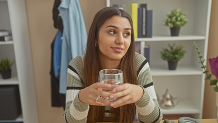 A thoughtful young hispanic woman holds a glass of water in her cozy home interior.