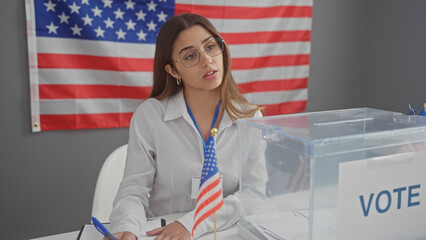 A focused young hispanic woman monitors a vote box indoors with an american flag backdrop,...