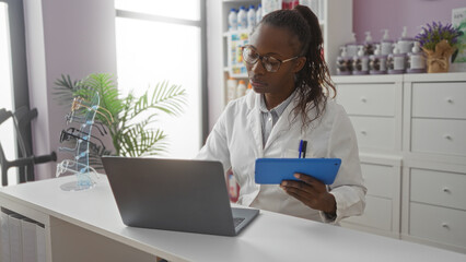 African american woman working in a pharmacy, wearing a white coat and glasses, using a laptop and tablet at the counter with eyeglasses display and pharmacy items in the background.