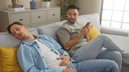 Two men, a loving couple, relax together at home, with one man sleeping on the sofa while the other checks his phone, showcasing a calm, intimate moment in a modern living room.