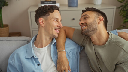 Fototapeta premium Two men sitting together in a cozy living room, smiling and gazing at each other, depicting a loving gay couple enjoying a moment of affection in their home.