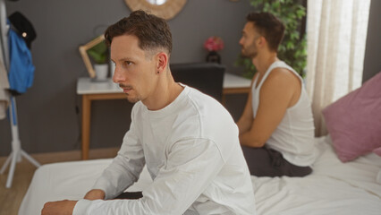 Fototapeta premium Two men sitting on a bed in a bedroom, one looking thoughtful while the other sits in the background, suggesting a moment of contemplation or discussion in their home environment